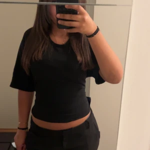 Svart croppad topp med knyt i ryggen - Säljer en svart croppad topp med korta ärmar och snygg knytning i ryggen som ger en öppen rygg-effekt. Toppen har en enkel och stilren design, perfekt att matcha med både jeans och kjol. Materialet känns mjukt och följsamt mot huden.