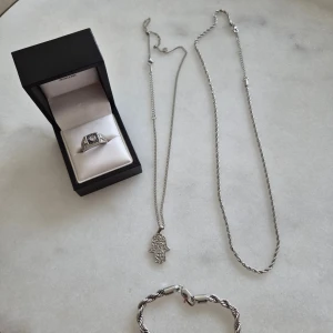 Smyckeset i rostfrittstål - Säljer ett snyggt smyckeset i silverfärg som innehåller en ring med svart och vit detalj samt två halsband, varav ett med hamsa-hand hänge och ett tvinnat kedjehalsband. Dessutom ingår ett matchande armband i tvinnad design. Perfekt för dig som gillar stilrena accessoarer. Rostfrittstål går att duscha med och tål svett.