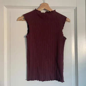 Ribbad vinröd ärmlös topp från Lindex - Vinröd ärmlös topp från Lindex i ribbad ekologisk bomull. Toppen har en hög hals med vågig kant och samma vågiga detalj nertill. 