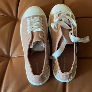 Rosa sneakers med pärldetaljer Anna Field - Säljer ett par ljusrosa sneakers från Anna Field med vita pärldetaljer och prickar på sidorna. Skorna har breda, glansiga satinsnören och vit sula samt tåparti. Perfekta för dig som vill ha något unikt och snyggt till vardagslooken.