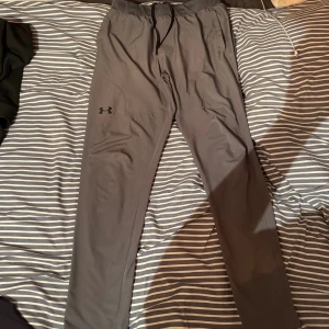 Grå träningsbyxor från Under Armour - Snygga grå träningsbyxor från Under Armour med justerbar midja och dragsko. Byxorna har en smal passform och är tillverkade i ett mjukt och stretchigt syntetmaterial. Perfekta för gymmet eller löprundan, med diskret logga på benet.