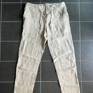 Beiga linnebyxor från These Glory Days - Snygga beiga linnebyxor från These Glory Days i storlek 46. Byxorna har dragsko i midjan, raka ben och är tillverkade i en skön mix av linne och polyester. Perfekta för varma dagar när du vill ha en avslappnad men stilren look.