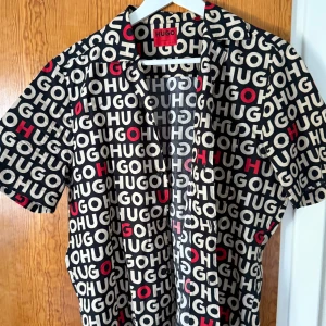 Svart kortärmad skjorta från Hugo - Säljer en svart kortärmad skjorta från Hugo med stort, grafiskt logomönster i vitt och rött över hela plagget. Skjortan har klassisk krage, knäppning framtill och är tillverkad i mjuk bomull. Perfekt för dig som vill sticka ut med en cool statement-look.