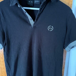 Svart pikétröja från Armani Exchange - Svart pikétröja från Armani Exchange med korta ärmar och klassisk krage. Tröjan har knappar framtill med randiga detaljer i grått och vitt samt broderad logga på bröstet. Tillverkad i bomull för en skön och avslappnad känsla.