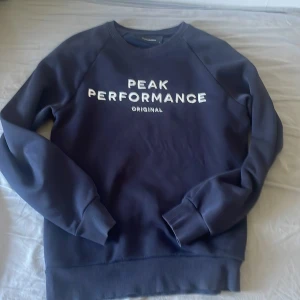 Mörkblå Peak Performance sweatshirt - Säljer en mörkblå sweatshirt från Peak Performance med vit logga framtill. Tröjan har rund hals, ribbade muddar och är tillverkad i mjuk bomullsmix. Perfekt för en chill och sportig stil.