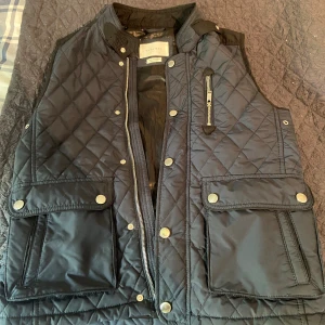 Quiltad marinblå väst från Zara Man - Snygg quiltad marinblå väst från Zara Man med silverfärgade knappar och dragkedjor. Västen har flera praktiska fickor framtill och hög krage. Perfekt för lager-på-lager och ger en clean look till din outfit. 