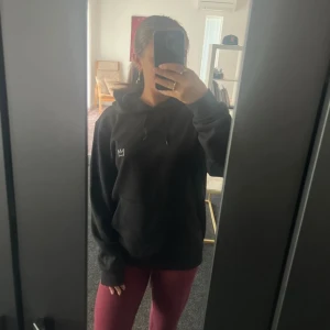 Svart Hov1 hoodie med turnétryck - Svart hoodie från Hov1 med broderad krona fram och stort turnétryck på ryggen med texten 'Vi Dansar Vidare I Livet'. Klassisk känguruficka, snörning i huvan och mjukt material. Perfekt för dig som älskar svensk musik och streetstyle.