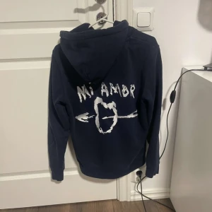 Mörkblå hoodie Mira Paris Mi Amor - Mörkblå hoodie från Mira Paris med huva och snörning. På ryggen finns ett vitt tryck med texten 'Mi Amor' och ett hjärta med pilar. Klassisk känguruficka framtill och mjukt material som passar perfekt till chill dagar.
