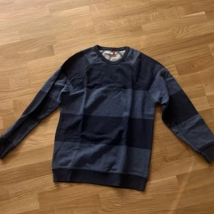 Randig blå sweatshirt från Homebound - Mörkblå och ljusblå randig sweatshirt från Homebound. Tröjan har rund halsringning, långa ärmar och breda ribbade muddar vid ärmslut och nederkant. Perfekt för en chill och stilren look.