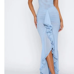 Ljusblå långklänning med volang - Supersnygg ljusblå långklänning med axelbandslös design och volangdetaljer längs slitsen och upptill. Klänningen har en figurnära passform med rynkade partier och är tillverkad i ett mjukt, lätt material som ger en elegant siluett. Perfekt för dig som vill sticka ut med en unik look.