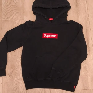Svart Supreme hoodie med box logo - Supreme hoodie i svart med ikonisk röd box logo på bröstet. Klassisk huva med snörning och ribbade muddar. Tillverkad i 100% bomull i storlek M📏. Perfekt för dig som gillar streetwear och vill sticka ut. Använd i fint skick⭐