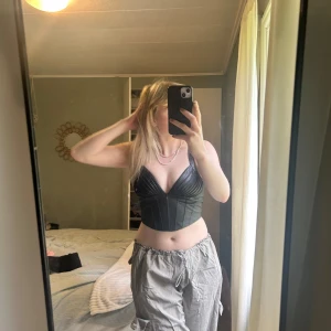 Svart korsett-topp från Amisu - Säljer en svart korsett-topp från Amisu i snyggt skinnliknande material. Toppen har breda axelband, djup v-ringning och coola sydda detaljer över bysten. Dragkedja i sidan för enkel påklädning. Perfekt för dig som vill sticka ut med en edgy look.