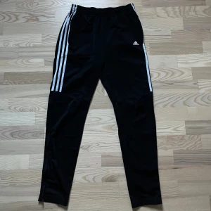 Svarta Adidas träningsbyxor - Svarta träningsbyxor från Adidas med klassiska vita ränder längs sidorna och logga på låret. Byxorna har en slim passform och är gjorda i mjukt syntetmaterial som andas bra. Perfekta för gymmet eller sportaktiviteter.