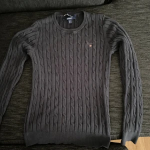 Mörkgrå kabelstickad tröja från GANT - Snygg mörkgrå kabelstickad tröja från GANT i stretchig bomull. Klassisk rund halsringning och diskret logga på bröstet. Långärmad modell med ribbade muddar och smal passform. Perfekt för lager-på-lager och stilrena outfits.