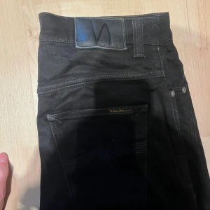 Svarta  jeans från Nudie Jeans - Säljer ett par svarta jeans från Nudie Jeans i storlek W32 L34. Jeansen har klassisk femficksdesign, svarta sömmar och Nudie-logga på bakfickan. Snygg passform med smala ben och tillverkade i mjukt bomullstyg med stretch.