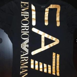 Äkta Emporio Armani (EA7) - Svart t-shirt från EA7 Emporio Armani i storlek XXL med stora, glansiga guldfärgade tryck på både fram- och baksida. Snyggt grafiskt mönster och logga som sticker ut. Rund hals och korta ärmar, tillverkad i mjuk bomull för skön känsla. Säljes pga att jag inte använder den så mycket. 