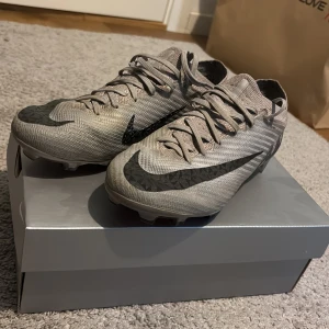 Nike Zoom Vapor 15 elits FG AS fotbollsskor grå/svart - Snygga Nike Zoom fotbollsskor i grått med svarta detaljer och stora Swoosh-loggor. Skorna har snörning, platt sula med dobbar och rosa detaljer undertill. Ovandelen är i syntetmaterial för lätt känsla och bra bollkontroll. Perfekta för dig som vill sticka ut på planen. Nypris ca 3100kr. Pris är inte hugget i sten.