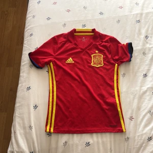 Spaniens landslagströja Adidas 140 - Röd fotbollströja från Adidas för Spaniens landslag med gula detaljer och broderat klubbmärke på bröstet. Tröjan har korta ärmar, V-ringning och gula ränder längs sidorna. Tillverkad i lätt och ventilerande ClimaCool-material. Har aldrig använt!💞