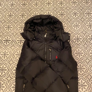 Svart dunjacka från Polo Ralph Lauren - Svart dunjacka från Polo Ralph Lauren med huva och dragkedjor framtill. Jackan har en klassisk puffer-design, röd broderad logga på bröstet och flera praktiska fickor med dragkedja. Perfekt för kalla dagar och riktigt snygg, har bara använt jackan under förra vinter