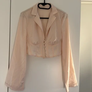 Champagnefärgad croppad kavaj Boohoo - Snygg croppad kavaj från Boohoo i champagnefärgad satin. Jackan har lång ärm, markerad midja och knappar framtill. Klassisk krage och figurnära passform som ger en trendig siluett. Perfekt för dig som vill sticka ut med en chic look. Aldrig använd