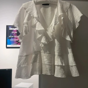 Vit volangblus från Zara - Supersöt vit blus från Zara med v-ringning och stora volanger längs halsringningen och ärmarna. Blusen har korta ärmar och är tillverkad i ett lätt, krinklat tyg som ger en luftig känsla. Perfekt för dig som gillar romantiska detaljer och flowy vibes. M men passar S