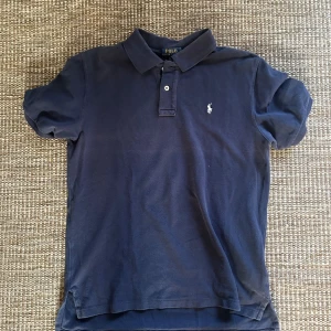 Marinblå pikétröja från Polo Ralph Lauren - Klassisk marinblå pikétröja från Polo Ralph Lauren med korta ärmar och krage. Tröjan har två knappar framtill och den ikoniska broderade loggan på bröstet. Tillverkad i mjuk bomull som är skön att bära hela dagen.