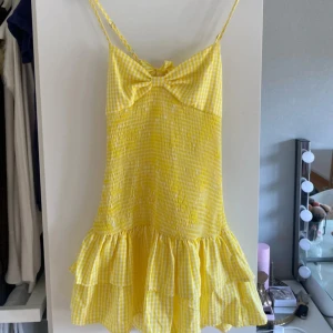 Gul rutig klänning med volang - Supergullig gul och vit rutig kortklänning med smala axelband och stor rosett framtill. 