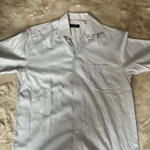 Ljusblå kortärmad skjorta från Eton - Säljer en stilren ljusblå skjorta från Eton med korta ärmar och klassisk krage. Skjortan har diskreta små prickar i tyget, en bröstficka och knäppning framtill. Perfekt för dig som gillar en clean och klassisk look.