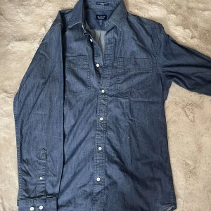 Blå chambrayskjorta från Gant S - Säljer en snygg blå chambrayskjorta från Gant Beacons Project i storlek S. Skjortan har två bröstfickor, vita knappar och klassisk krage. Regular fit och tillverkad i bomull. Perfekt för dig som gillar stilren och avslappnad look.