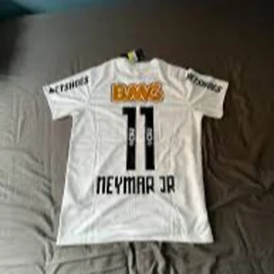 Vit Santos Neymar Jr fotbollströja Nike - Santos FC fotbollströja i vitt med Neymar Jr och nummer 11 på ryggen. Tröjan har korta ärmar, svarta tryck och flera sponsorloggor, bland annat BMG i orange på bröstet. Klassisk passform och tillverkad i lätt, ventilerande material.
