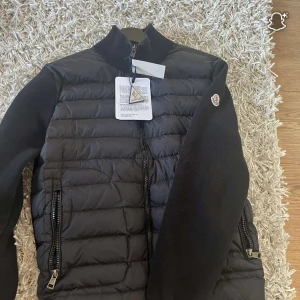 Svart Moncler pufferjacka med stickade ärmar - Snygg svart pufferjacka från Moncler med quiltad framsida och stickade ärmar. Jackan har dragkedja framtill, två dragkedjefickor och Moncler-logga på ärmen. Perfekt för dig som vill ha en stilren och exklusiv look.