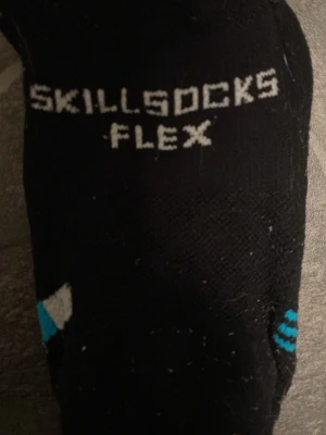 Svarta Skillsocks Flex med grepp - Säljer ett par svarta Skillsocks Flex med vita grepp på undersidan och blå detaljer. Perfekta för dig som vill ha extra fäste vid träning eller fotboll. Strumporna är höga och har en sportig look med logga upptill och på foten.