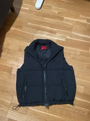 Svart dunväst från Hugo - Säljer en svart dunväst från Hugo med hög krage och två dragkedjeförsedda fickor. Västen har en diskret Hugo-logga på bröstet och justerbar dragsko nedtill. Perfekt för lager-på-lager.