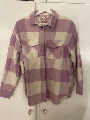 Rutig overshirt från H&M i lila och vanilj vitt - Säljer en oversized rutig overshirt från H&M i lila och beige. Jackan har stora bröstfickor med lock, klassisk krage och knäppning framtill. Perfekt för lager på lager och ger en chill vibe till din outfit.