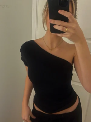 Svart oneshoulder topp från Zara - Säljer en svart oneshoulder topp från Zara i ribbat material. Toppen har en tight passform och är croppad, vilket ger en snygg siluett. Perfekt för dig som vill ha en enkel men stilren topp med unik skärning.