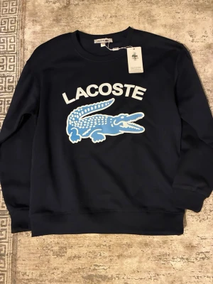 Lacoste sweater -  En blå Lacoste sweatshirt är en klassisk tröja gjord i mjuk bomullsfleece. Den haren rund hals och den ikoniska krokodil-loggan broderad på framsidan av tröjan. Perfekt för en avslappnad men stilren look.