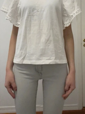 Vit blus med volangärmar från Kappahl  - Säljer en söt vit blus med breda volangärmar och dekorativ hålspets längs ärmsluten. Blusen har en rak passform och är tillverkad i ett lätt bomullstyg som känns luftigt och fräscht. Perfekt till jeans eller kjol för en enkel men fin look.