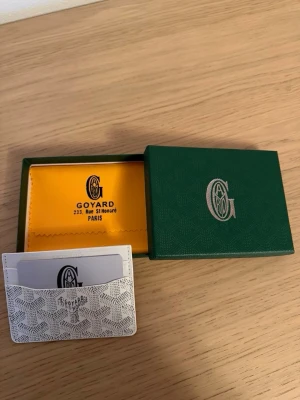 Vit kortfodral  - Säljer ett stilrent vitt kortfodral med ikoniskt mönster och flera fack för kort. Tillverkad i exklusivt skinn och levereras med original ask och dustbag. Perfekt för dig som vill ha en lyxig och smidig accessoar i fickan.