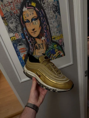 Nike Air Max 97 Gold Metallic - Nike Air Max 97 i metallic guld med vita och svarta detaljer samt röd swoosh. Skon har den ikoniska vågiga designen, synlig Air-sula och snörning. Perfekt för dig som vill sticka ut med en klassisk sneaker i unik färg.