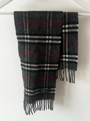 Rutig halsduk från Burberry i ull - Klassisk halsduk från Burberrys i mörkgrått, svart, vitt och rött. Rutigt mönster och fransar i ändarna. Tillverkad i 100% lammull och har en snygg vintage-vibe. Perfekt accessoar för kalla dagar.