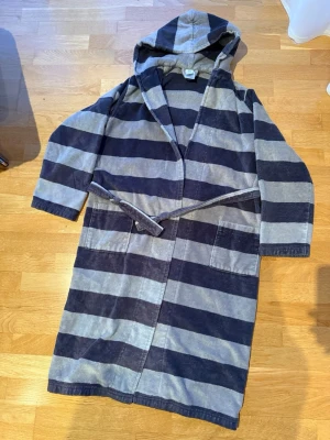 Randig frottébadrock från Sanetta - Mysig badrock från Sanetta i storlek 164. Badrocken har breda mörkgrå och ljusgrå ränder, huva, två stora fickor och knytskärp i midjan. Tillverkad i mjuk frotté som är perfekt efter duschen eller badet.