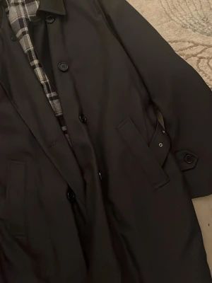 Svart rock med rutigt foder - Snygg svart rock med klassisk krage och knäppning framtill. Insidan har ett stilrent rutigt foder i svart, vitt och grått. Två fickor med lock på sidorna och diskreta knappar. Perfekt för dig som vill ha en clean och tidlös look. Vatten tålig och bara använd en gång