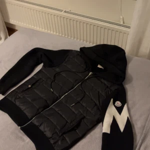 Svart pufferjacka med huva från Moncler - Säljer en svart pufferjacka från Moncler med huva och dragkedja. Jackan har stickade ärmar, quiltad kropp och coolt blixtmönster i vitt på ena ärmen. Två fickor framtill och snörning i huvan. Perfekt för dig som vill ha en streetig look.