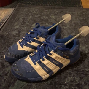 Blåvita Adidas Stabil sneakers - Säljer ett par blå och vita Adidas Stabil sneakers med klassiska tre ränder på sidorna. Skorna har snörning, meshpartier för andning och robust gummisula i brunt. Perfekta för dig som gillar sportig stil och vill ha bekväma skor med bra grepp. Storlek 42