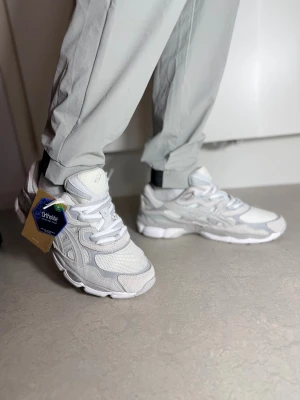 Vita Asics sneakers med mesh och mocka snören  - Fräscha Asics sneakers i ljusgrått och vitt med snygga detaljer i mesh och mocka. Skorna har rund tå, platt sula och snörning. Ortholite komfortsula för extra skön känsla. Perfekt för dig som gillar stilrena och sportiga sneakers. Dubbla snören. Storlek  44 finns. 