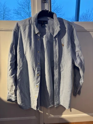 Ljusblå skjorta från Ralph Lauren - Klassisk ljusblå skjorta från Ralph Lauren med ikonisk broderad logga på bröstet. Skjortan har knappar hela vägen, krage och långa ärmar. Tillverkad i mjuk bomull som känns skön mot huden. Perfekt till jeans eller chinos för en clean look.