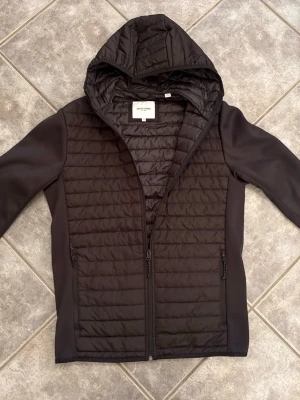 Svart quiltad hybridjacka från Jack & Jones - Säljer en svart quiltad jacka från Jack & Jones med huva och dragkedja. Jackan har släta ärmar i kontrastmaterial och två fickor med dragkedja. Perfekt som övergångs/vårjacka. Använd fåtal gånger. Aldrig tvättad.