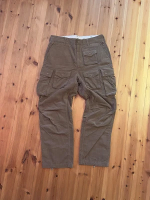 Bruna cargobyxor från Engineered Garments - Säljer ett par bruna cargobyxor från Engineered Garments. Byxorna har flera stora fickor med lock och knapp, raka ben och är tillverkade i ett slitstarkt bomullstyg. Perfekta för dig som gillar funktionella och snygga byxor med streetwear-vibe.