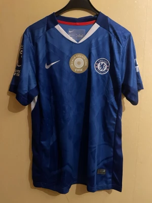 Chelsea hemmatröja – João Pedro #20 – FIFA World Champions - en sprillans ny Chelsea hemmatröja i storlek M, helt oanvänd med tags kvar. Klassisk blå design med diskret mönster, Chelsea-emblem på bröstet samt FIFA World Champions-märke. Tröjan har Premier League-patch och ”No Room For Racism”-märke på ärmen. Tryck på ryggen: JOÃO PEDRO 20. Tillverkad i Nike Dri-Fit-material som andas och ger hög komfort. Perfekt för match, träning eller samling. I absolut nyskick.