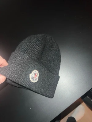 Svart Moncler mössa i ull - Säljer en svart ribbstickad mössa från Moncler med klassisk logga framtill. Mössan är i mjuk ull och har vikt kant för extra stilpoäng. Perfekt accessoar för kalla dagar och snygg till alla outfits.
